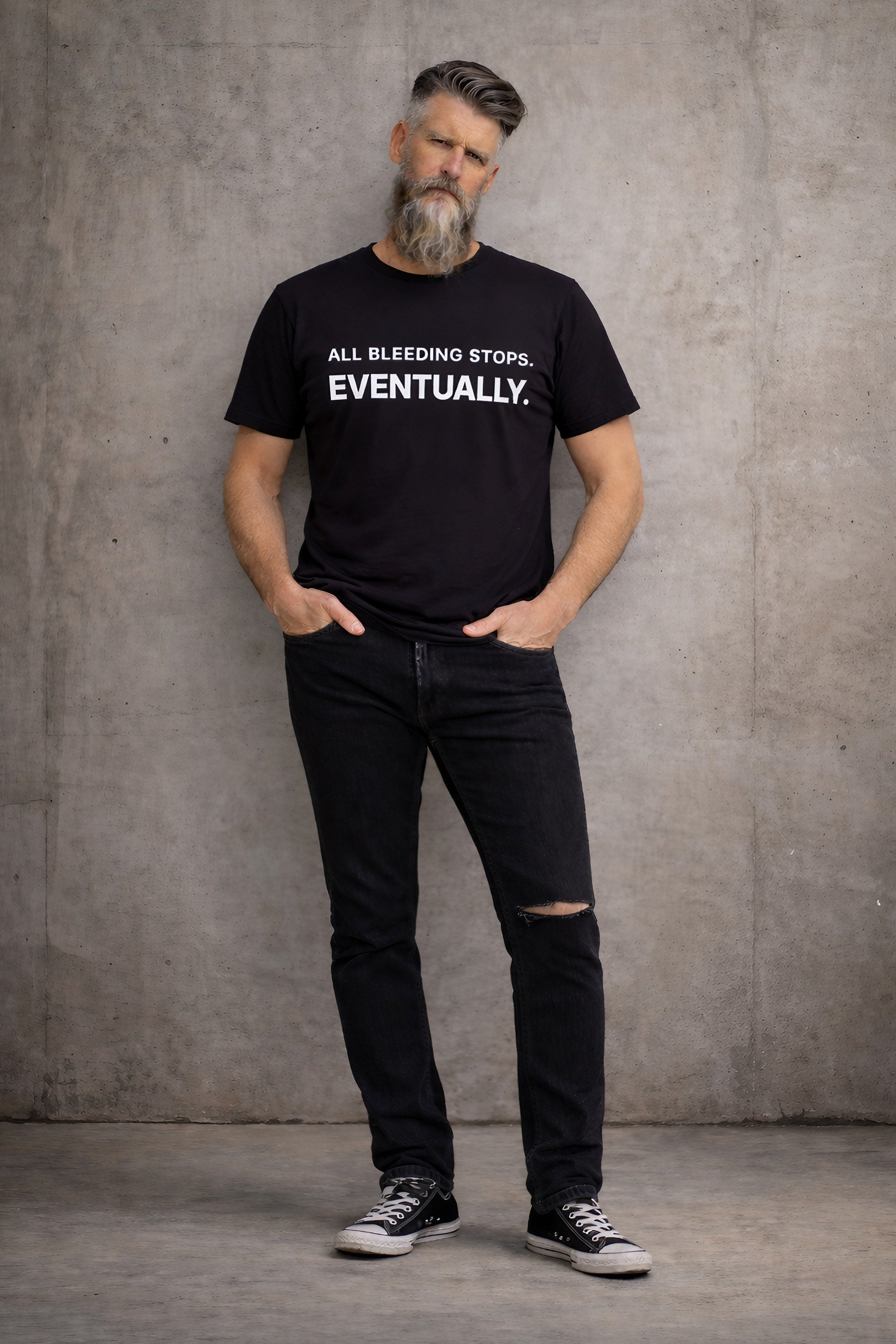 Man wearing black “All Bleeding Stops Eventually” Bitter Bull graphic t-shirt standing against raw concrete wall, full body, minimal streetwear style