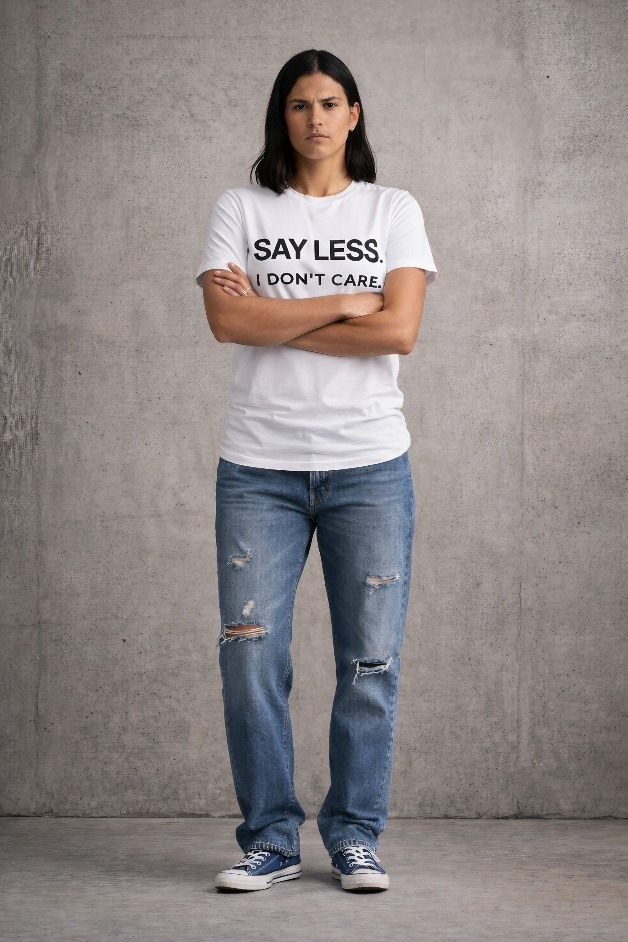 Woman wearing white Bitter Bull graphic t-shirt with ‘Say Less I Don’t Care’ text, arms crossed, full body model against concrete wall